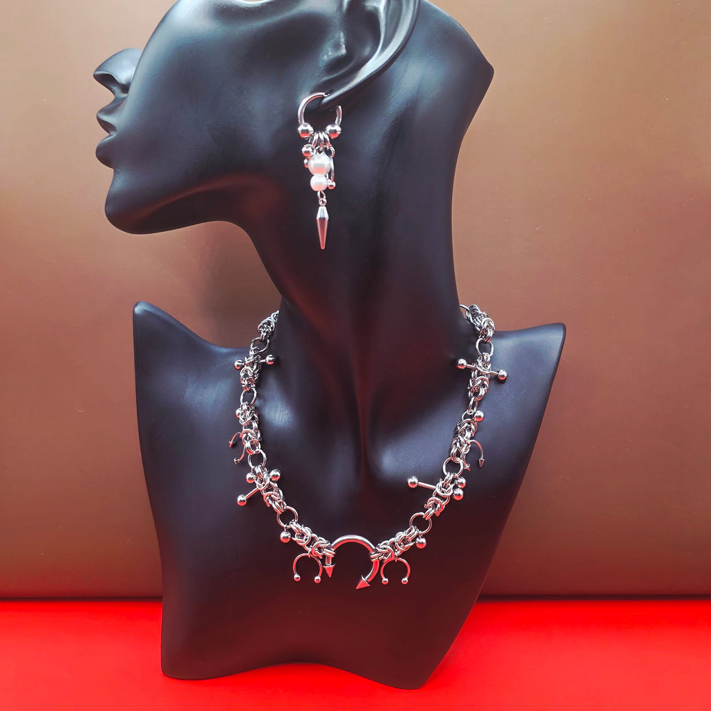 A silver-toned jewelry set displayed on a black mannequin bust, featuring a matching necklace and earring with pink bead accents, set against a brown background with a red surface.