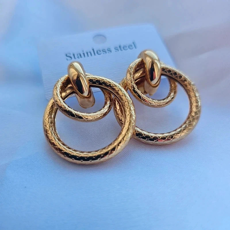 A pair of gold-toned double-ring hoop earrings with a textured, snake-like surface finish, displayed on a white backing card labeled 'Stainless steel' against a soft light blue background.