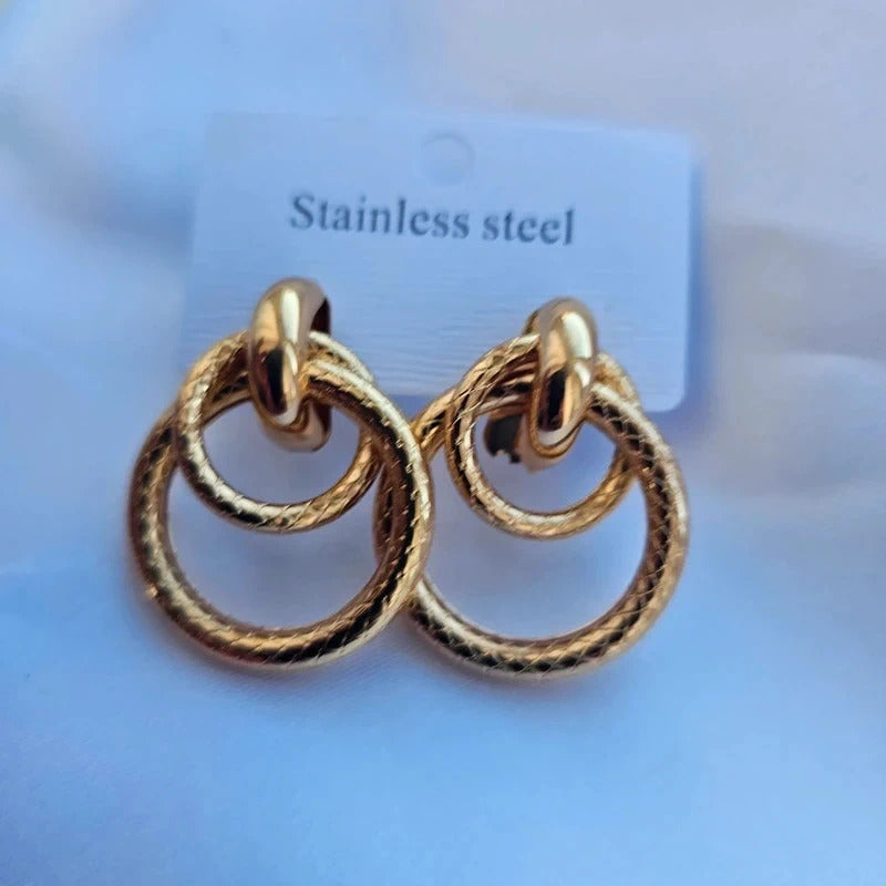 A pair of gold-toned stainless steel hoop earrings with a textured rope-like finish and a double-ring design, displayed on a white backing card labeled 'Stainless steel' against a light blue background.
